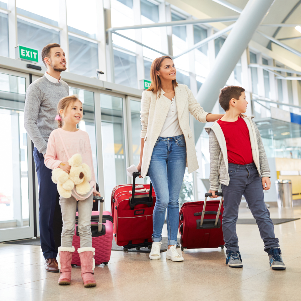 High-quality, professional photograph of a multiracial family of four (mother, father, and two children) smiling and walking hand-in-hand along a scenic tropical beach at sunset, with light waves rolling in. The image visually represents the joy and peace of a well-planned family trip managed by a specialized tour and travel agency for a family trip.